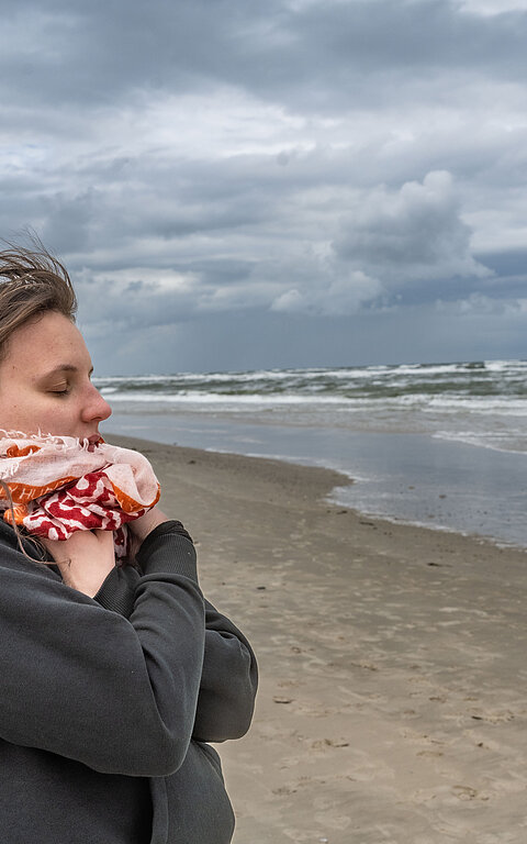 Frau an der Wasserkante am Strand von Juist im Herbst oder Winter, atmet tief ein und vergräbt das Kinn in einem bunten Schal, ihre Haare fliegen im Wind
