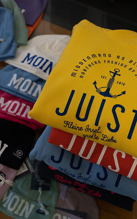 Typische Produkte für Juist mit erkennbarer Aufschrift kaufen, bunte Pullover aufgestapelt mit der Aufschrift "Juist", da neben Mützen mit der Aufschrift "Moin", ebenfalls bunt