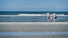 Familie an der Wasserkante der Nordsee auf Juist, zwei Frauen, vermutlich Oma und Mutter, halten ein Kind zwischen sich an den Händen