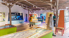 Ausstellung innen im Nationalpark-Haus auf Juist, Sicht von der Kasse und dem Eingang aus auf die bunten Exponate, Frau steht an einer der Stationen - Person ist unscharf und läuft durch das Bild