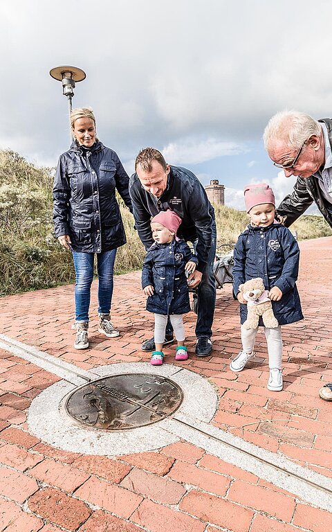 Familie auf der Juister Strandpromenade im Sonnenschein schaut sich die Markierung des 7. Längengrads an