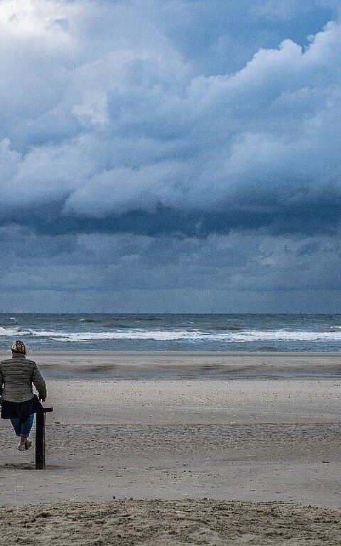 Wasserkante der Nordsee auf Juist am Strand mit blick auf zwei Personen auf einer Bank und bewölktem Himmel